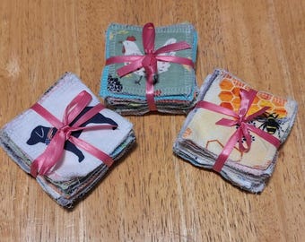 May include: Three stacks of square fabric cloths tied with pink satin ribbons. One stack features a black dog silhouette, another displays a white chicken on a green background, and the third shows a yellow honeycomb pattern with a bee illustration.