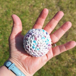 May include: A small, round, crocheted ball in a hand. The ball is primarily white with flecks of purple, blue, and pink. The ball is held in an open palm, with a light blue watch band visible in the lower left corner.