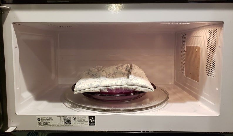 A filled reusable microwave potato bag on a microwavable plate inside a microwave oven in the color of white with light blue floral design. This product is a sewing tutorial to make potato bags.