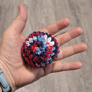 May include: A small, round, crocheted ball in shades of red, white, and blue. The ball is made of yarn and appears to be handmade. The ball is held in an open hand, showcasing the texture and color variations of the yarn.