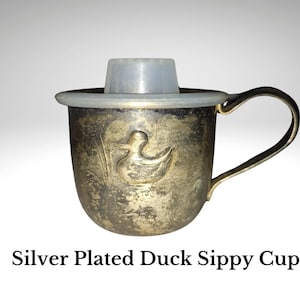 May include: Silver-plated sippy cup with a duck design. The cup has a handle and a plastic insert. The text "Silver Plated Duck Sippy Cup" is at the bottom of the image.