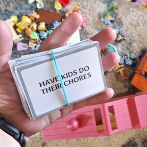 May include: A stack of white cards held together with a teal rubber band. The cards have the text "HAVE KIDS DO THEIR CHORES" in black. The background includes toys and a pink toy house.