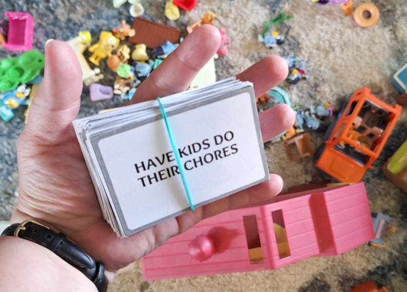 May include: A stack of white cards held together with a teal rubber band. The cards have the text "HAVE KIDS DO THEIR CHORES" in black. The background includes toys and a pink toy house.