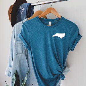 Puede incluir: Camiseta de color turquesa con un contorno blanco de la forma del estado de Carolina del Norte. La camiseta est&aacute; colgada en una percha de madera, junto con una camisa azul claro y un sombrero marr&oacute;n en el fondo. La camiseta es de material suave.
