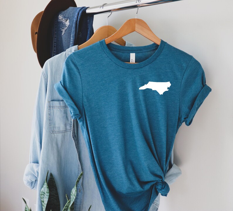 Puede incluir: Camiseta de color turquesa con un contorno blanco de la forma del estado de Carolina del Norte. La camiseta est&aacute; colgada en una percha de madera, junto con una camisa azul claro y un sombrero marr&oacute;n en el fondo. La camiseta es de material suave.