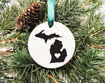 May include: White ceramic ornament with a black silhouette of the state of Michigan with a small white heart in the center. The ornament is hanging from a green ribbon.