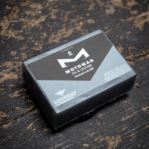 Może przedstawiać: Szare mydło z napisem "MOTOMAN OIL & LEATHER Handmade in the USA" na etykiecie. Mydło leży na ciemnym drewnianym podłożu.
