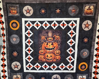 Könnte beinhalten: Ein Halloween-Quilt mit einem zentralen Bild von grinsenden Kürbissen. Der Quilt hat einen schwarzen Hintergrund mit orangefarbenen und weißen Akzenten, Sternen und den Worten "HAPPY HALLOWEEN" in bunten Buchstaben. Der Rand hat ein sich wiederholendes Rautenmuster.