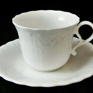 May include: White porcelain teacup and saucer set with a delicate floral design. The cup has a rounded shape and a handle, while the saucer has a scalloped edge.