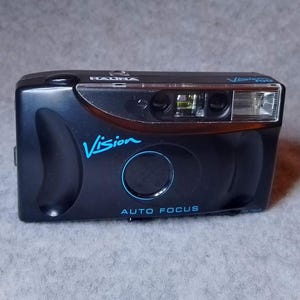 May include: A black compact camera with a blue "Vision" logo and "Auto Focus" text. The camera has a built-in flash and a lens cover.