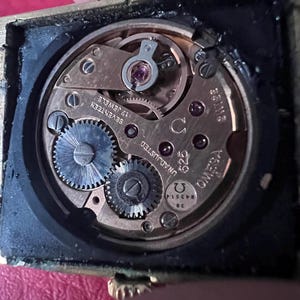 May include: Close-up of a gold-toned watch movement with the text "OMEGA" and "UNADJUSTED" visible. The movement has 17 jewels and is labelled "SEVENTEEN".