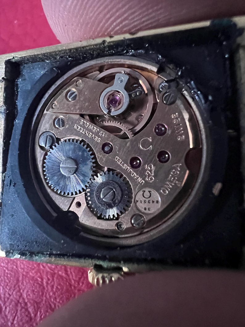 May include: Close-up of a gold-toned watch movement with the text "OMEGA" and "UNADJUSTED" visible. The movement has 17 jewels and is labelled "SEVENTEEN".