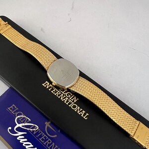 Puede incluir: Un reloj Elgin International dorado con una correa de malla, apoyado en una caja negra con letras doradas. El reloj tiene una esfera redonda y una parte trasera de acero inoxidable. La caja tambi&eacute;n tiene una secci&oacute;n azul con la palabra "Garant&iacute;a".
