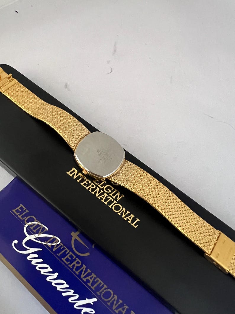 Puede incluir: Un reloj Elgin International dorado con una correa de malla, apoyado en una caja negra con letras doradas. El reloj tiene una esfera redonda y una parte trasera de acero inoxidable. La caja tambi&eacute;n tiene una secci&oacute;n azul con la palabra "Garant&iacute;a".