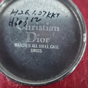 Puede incluir: Primer plano de la parte posterior de un reloj plateado con el texto "Christian Dior" y "Watches All Steel Case Swiss" impreso en &eacute;l. El reloj tambi&eacute;n tiene texto escrito a mano que dice "750 1.37 KKT H212".