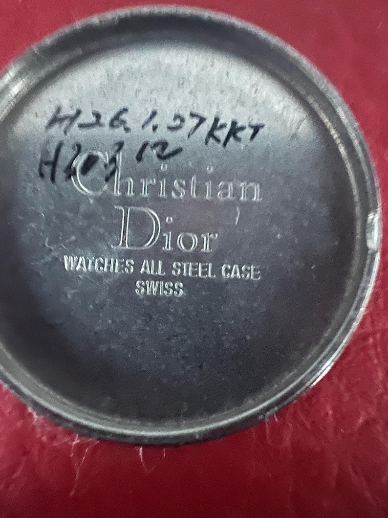 Puede incluir: Primer plano de la parte posterior de un reloj plateado con el texto "Christian Dior" y "Watches All Steel Case Swiss" impreso en &eacute;l. El reloj tambi&eacute;n tiene texto escrito a mano que dice "750 1.37 KKT H212".