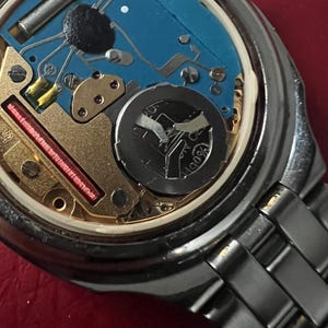 Puede incluir: Primer plano de la parte posterior de un reloj de pulsera plateado con un mecanismo interno azul y dorado. El reloj tiene una pulsera de metal plateado.