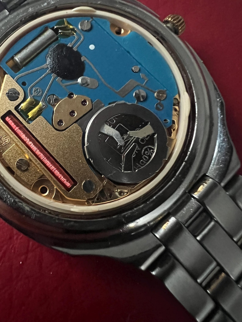 Puede incluir: Primer plano de la parte posterior de un reloj de pulsera plateado con un mecanismo interno azul y dorado. El reloj tiene una pulsera de metal plateado.