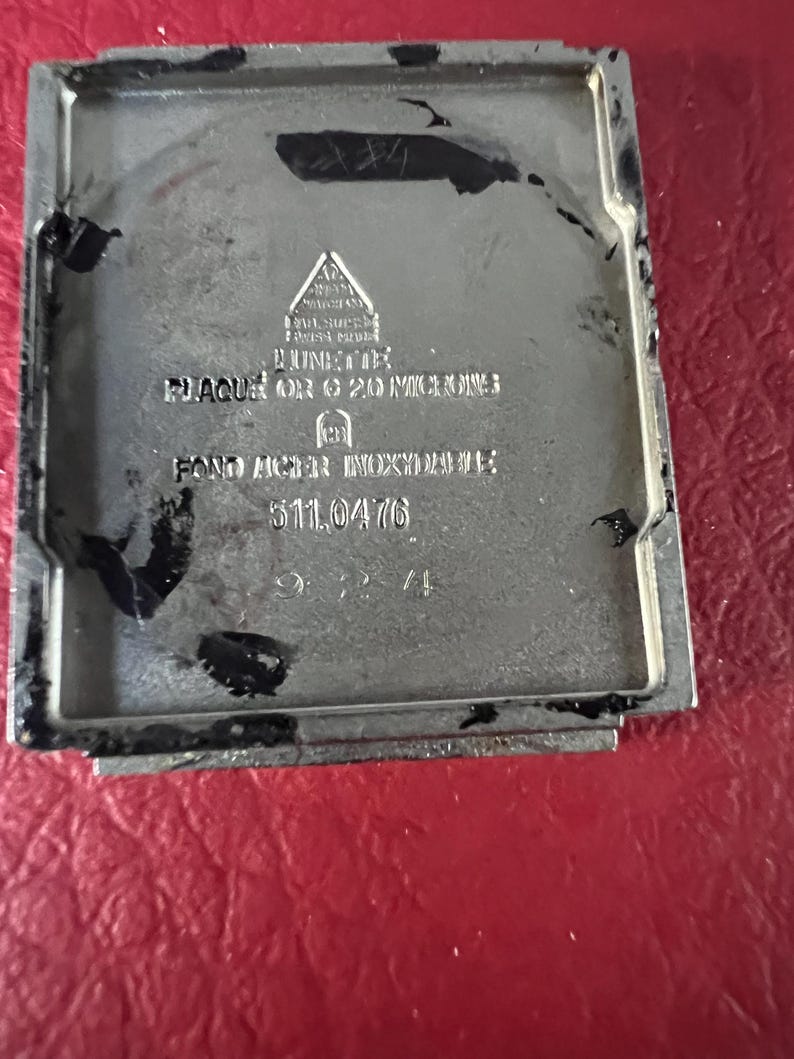 May include: A silver watch back with the text "PLAQUE OR 20 MICRONS FONDO ACER INOXIDABLE 511.0476"