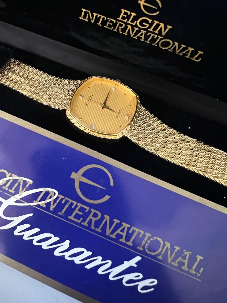 Puede incluir: Reloj Elgin International de cuarzo dorado con esfera texturizada y correa de malla. La esfera del reloj tiene detalles de diamantes y el nombre de la marca. El reloj se muestra en una caja negra con letras doradas y una tarjeta de garant&iacute;a azul.