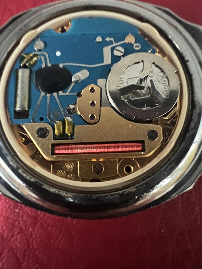 Puede incluir: Primer plano de la parte posterior de un reloj plateado con un mecanismo interno de color dorado. El reloj tiene un circuito impreso azul y una tapa de bater&iacute;a plateada con el texto "ROAMER" impreso en ella.