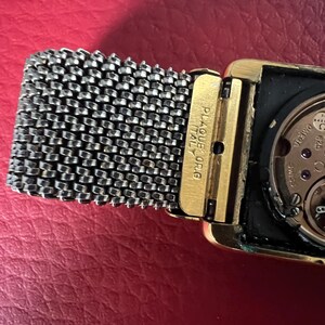 May include: A gold-toned wristwatch with a square face and a silver mesh band. The watch face is black and has a white dial with black hands. The watch has the text "PLAQUE OR ITALY" on the side.