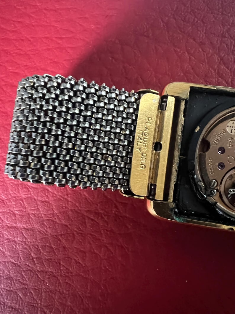 May include: A gold-toned wristwatch with a square face and a silver mesh band. The watch face is black and has a white dial with black hands. The watch has the text "PLAQUE OR ITALY" on the side.