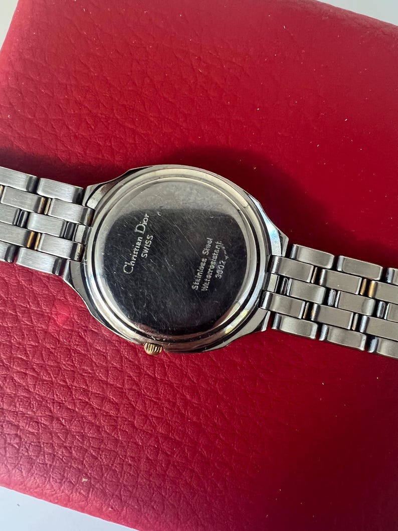 Puede incluir: La parte posterior de un reloj de pulsera de dos tonos, plateado y dorado, con una correa de acero inoxidable. La esfera del reloj tiene el texto "Christian Dior Swiss" y "Stainless Steel Waterresistant 30M/3ATM".