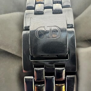 Puede incluir: Primer plano de una correa de reloj de plata y oro con un cierre que tiene las letras "CD" grabadas.