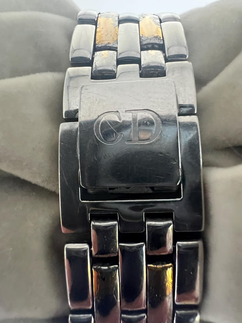 Puede incluir: Primer plano de una correa de reloj de plata y oro con un cierre que tiene las letras "CD" grabadas.