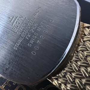 Puede incluir: Primer plano de la parte trasera de un reloj Elgin Quartz. La parte trasera de acero inoxidable tiene grabado: Elgin, Quartz, Stainless Steel Back, FK 266, Swiss MVT, y el n&uacute;mero de serie 0072904. Se ve una correa de reloj dorada y trenzada.