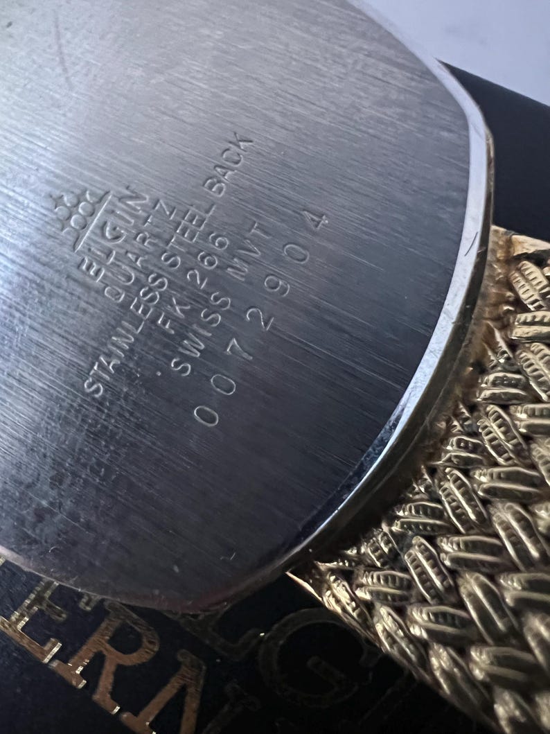 Puede incluir: Primer plano de la parte trasera de un reloj Elgin Quartz. La parte trasera de acero inoxidable tiene grabado: Elgin, Quartz, Stainless Steel Back, FK 266, Swiss MVT, y el n&uacute;mero de serie 0072904. Se ve una correa de reloj dorada y trenzada.