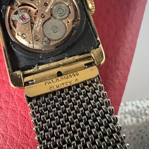 May include: A gold-tone wristwatch with a square face and a black mesh band. The watch face is visible and shows the inner workings of the watch. The watch has the text "PAT. N. 5128550 ELMITEX O" on the back.