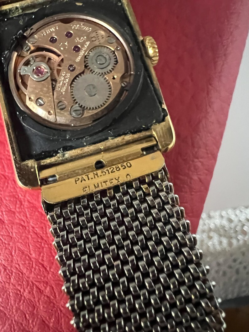 May include: A gold-tone wristwatch with a square face and a black mesh band. The watch face is visible and shows the inner workings of the watch. The watch has the text "PAT. N. 5128550 ELMITEX O" on the back.
