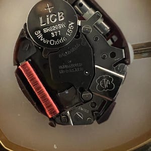 Puede incluir: Primer plano de un movimiento de reloj con una bater&iacute;a plateada etiquetada "LICB Silver Oxide SR626SW 377 1.55V". El movimiento tiene varios componentes met&aacute;licos y una bobina roja.