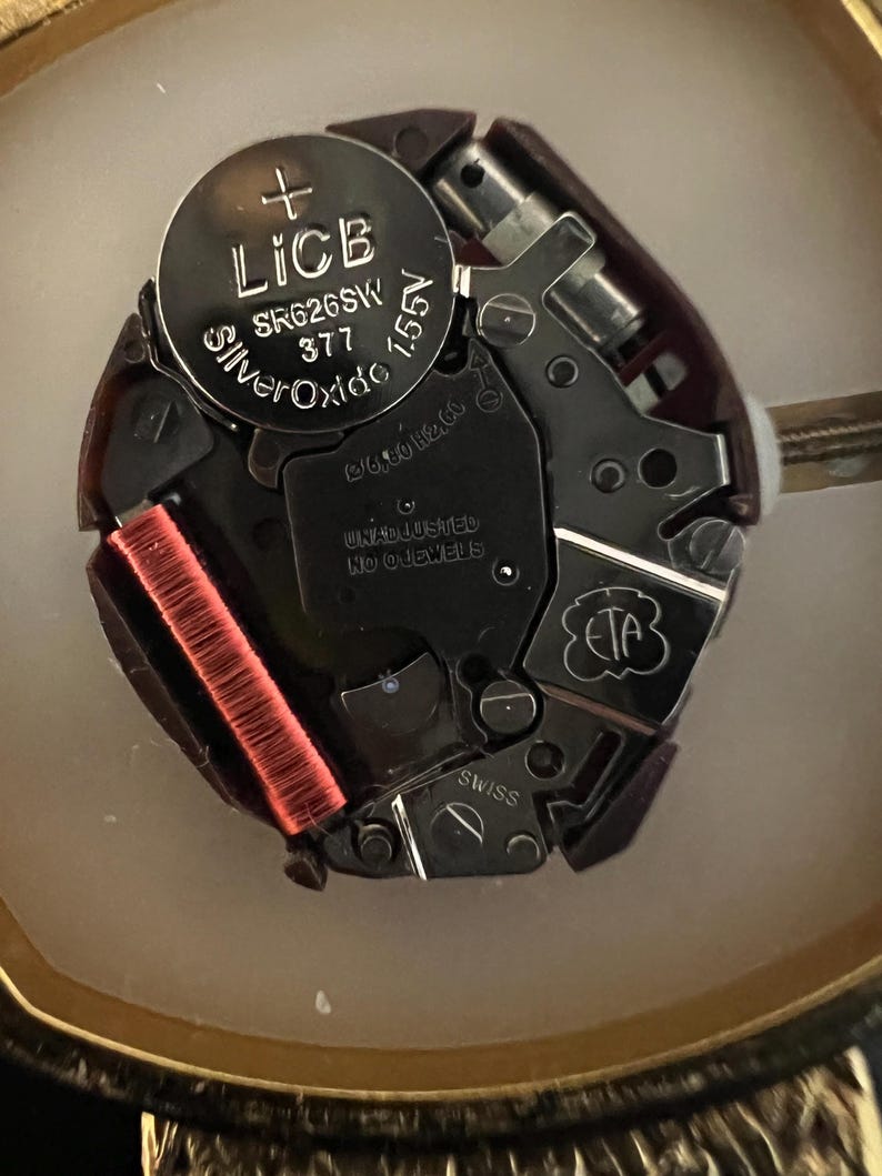 Puede incluir: Primer plano de un movimiento de reloj con una bater&iacute;a plateada etiquetada "LICB Silver Oxide SR626SW 377 1.55V". El movimiento tiene varios componentes met&aacute;licos y una bobina roja.