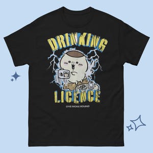 Puede incluir: Una camiseta negra de manga corta con un gráfico de un personaje de dibujos animados sosteniendo una tarjeta. El texto dice DRINKING LICENCE y ONE MORE ROUND en amarillo y azul. El diseño incluye rayos y una mesa pequeña con una bebida.