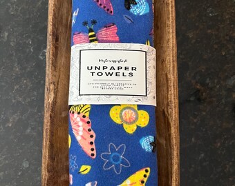 May include: A roll of blue fabric unpaper towels featuring a colorful pattern of butterflies and flowers. A white paper band wraps around the center with the text MyScrappyGirl UNPAPER TOWELS, eco-friendly alternative to paper towels, for best results, wash before using.