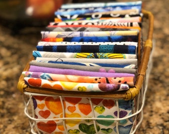 May include: A rectangular wire basket with a wooden rim containing a stack of folded fabric squares. The fabrics feature various patterns including multicolored hearts, floral designs, geometric shapes, and stripes in shades of blue, orange, and purple.