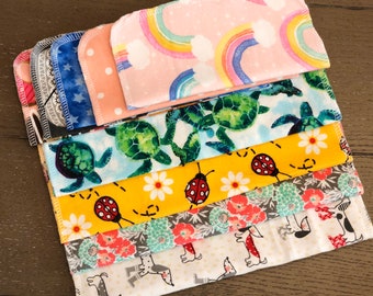 May include: A stack of rectangular fabric cloths featuring various patterns including rainbows on pink, green sea turtles on light blue, ladybugs and daisies on yellow, floral prints, and small dogs on a white background with stitched edges.