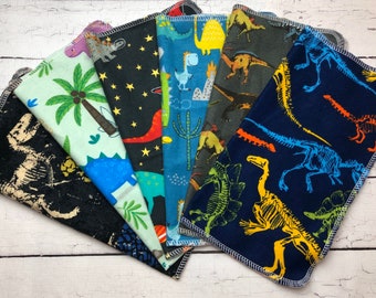 May include: A set of six fabric cloths featuring various dinosaur patterns. The designs include black with fossil skeletons, light green with palm trees, black with stars and dinosaurs, blue with cartoon dinosaurs, dark gray with dinosaurs, and navy with skeletons.