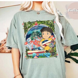 Può includere: T-shirt verde salvia con una grafica colorata del film d'animazione "Ponyo". Il design include il titolo del film, un personaggio maschile e vari elementi del film. La maglietta ha maniche corte e una vestibilità rilassata.