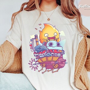May include: Cream-colored t-shirt featuring a colorful cartoon illustration of a moving castle with a fire demon on top. The design includes buildings, smoke, and mechanical elements. The word "Natural" is printed at the bottom.