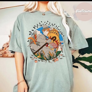 Può includere: T-shirt verde salvia con un gatto dei cartoni animati in abito, con un bastone e circondato da topi e pipistrelli. Il gatto è di fronte a un edificio con un ombrello a righe rosse e bianche. La maglietta ha una vestibilità ampia.