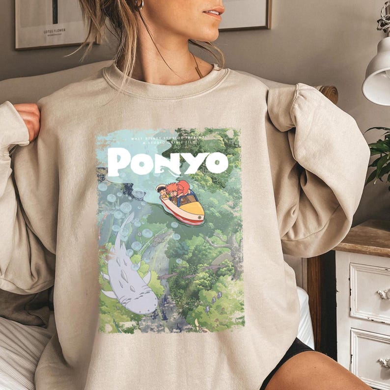 Puede incluir: Sudadera beige con un gr&aacute;fico de la pel&iacute;cula de animaci&oacute;n "Ponyo". El dise&ntilde;o incluye el t&iacute;tulo "Ponyo" en blanco, y una ilustraci&oacute;n de personajes en un barco y un pez grande. La sudadera es de color neutro.