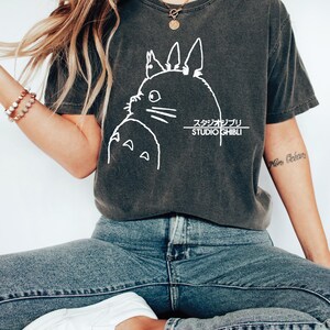 May include: Dark gray t-shirt featuring a white graphic of a cartoon character and Japanese text. The shirt is a crew neck and has short sleeves. The model is wearing blue jeans and white shoes.