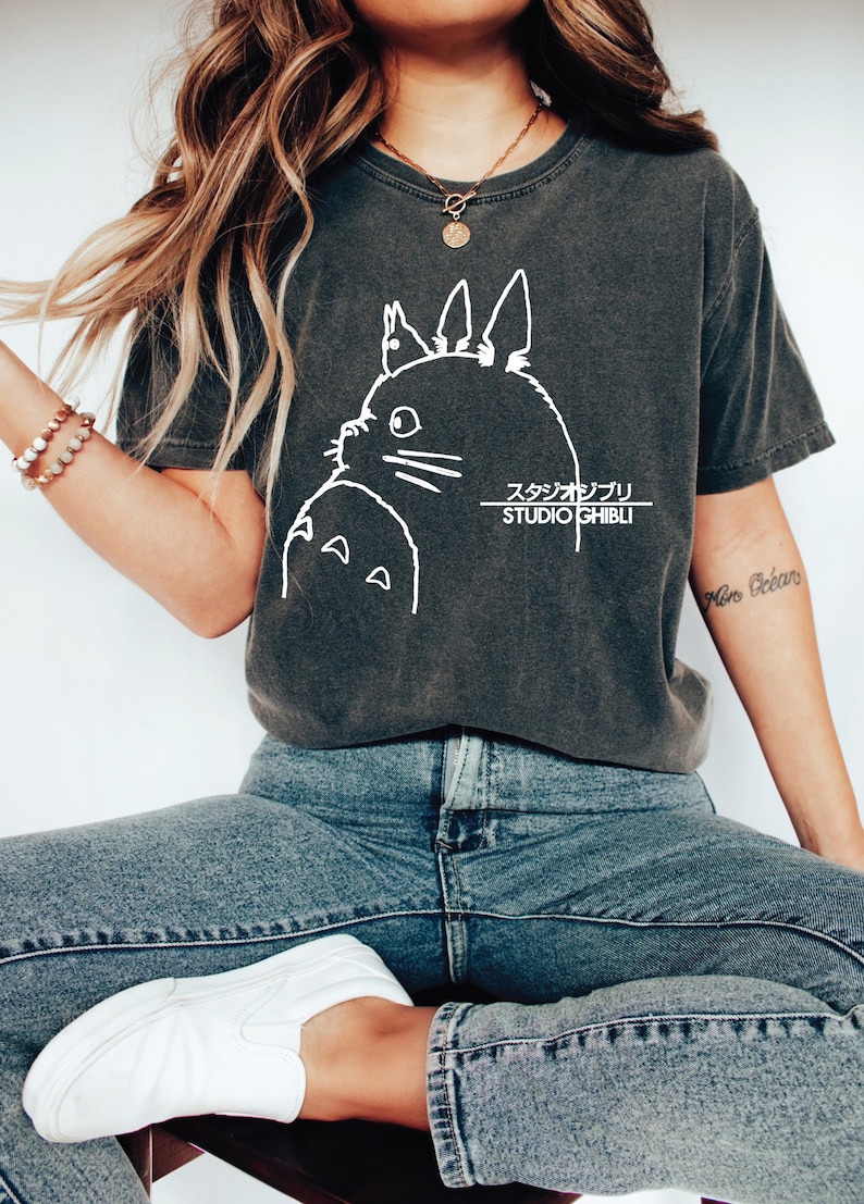 May include: Dark gray t-shirt featuring a white graphic of a cartoon character and Japanese text. The shirt is a crew neck and has short sleeves. The model is wearing blue jeans and white shoes.