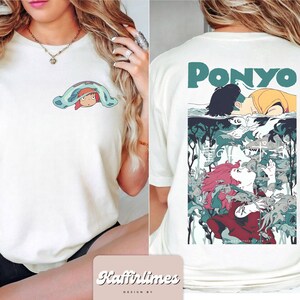May include: Cream-coloured t-shirt with a cartoon illustration from the film "Ponyo". The front displays a small graphic of a character under a blanket. The back features a larger graphic with the word "Ponyo" in teal.