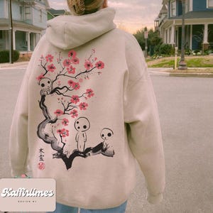 May include: Beige hoodie with a back graphic featuring a stylized cherry blossom branch with pink flowers and three small, white spirit figures. The hoodie has a drawstring hood and ribbed cuffs. The text "Kaffirlimes DESIGN BY" is at the bottom.