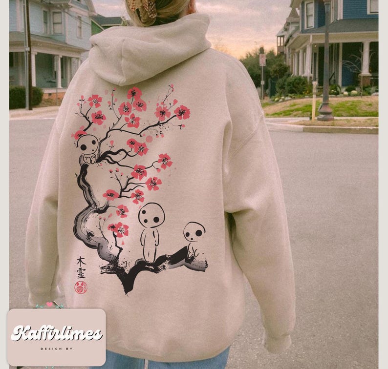 May include: Beige hoodie with a back graphic featuring a stylized cherry blossom branch with pink flowers and three small, white spirit figures. The hoodie has a drawstring hood and ribbed cuffs. The text "Kaffirlimes DESIGN BY" is at the bottom.